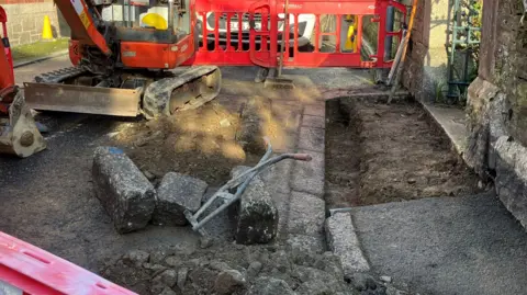 Excavated pavement in front of church gate with kerb stones pulled out and bare ground. Some tools and digger and bollards to prevent people walking there