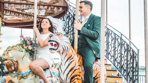 Felixine Hick Harmony and Seán smiling whilst riding a carousel in Cardiff Bay. Harmony, on the left, is wearing a short white wedding dress, a white vale and white mesh sleeves. She is holding a large bouquet of flowers. Sean, on the right, is wearing a dark green suit with a pale pink tie. 