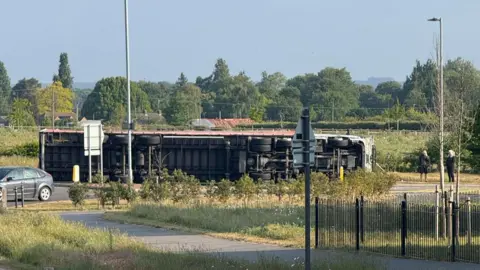 An overturned lorry rests on the side of a road. Its undercarriage can be seen. Another car drives nearby as people stand on the side of the road and watch on. 