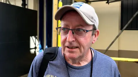 A middle-aged white man with glasses and a beige cap on looking slightly to his right. He has a black lanyard on and a black bag strap in his right shoulder.
