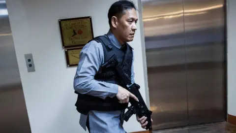 A man dressed in a blue shirt and a bullet-proof vest carrying a gun.