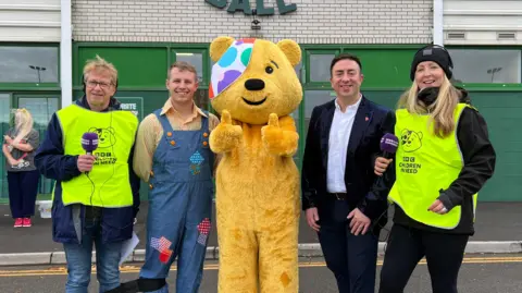 Four people are standing next to Pudsey bear at Huish Park.
