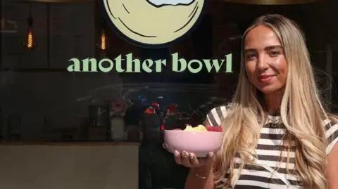 Contributed Chloe Marsh stands outside her cafe holding a pink bowl filled with fruit. She has long blonde hair that is lightly curled at the ends. She wears a striped black and white top.