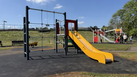 North Northamptonshire Council A newly upgraded children’s play area in Corby Old Village with modern, brightly coloured equipment. The playground includes level rubberised surfacing, wide pathways and play features set at different heights, including swings, climbing frames and interactive panels. The play area is surrounded by a low fence, with trees and nearby houses visible in the background on a clear day.
There is a group of people to the right. 