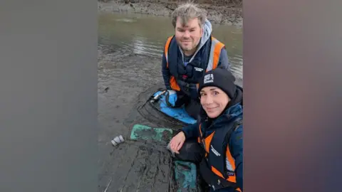 Durham Wildlife Trust Dr Martina Bristow and engagement officer Blair Watson take a selfie during seagrass planting at the River Don near Jarrow. They are wearing high visibility blue and orange vests as they sit in the mud next to a frame which holds the young seagrass plants. Dr Bristow has brown eyes and a pierced nose and lip. She is wearing a black beanie. Blair Watson has short grey hair and blue eyes.
