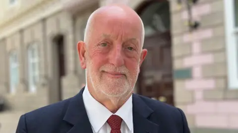 Deputy Steve Luce, a man with very short white hair on the side of his head and a white beard. He is wearing a white shirt, red and white spotty tie, and a dark blue blazer. He is smiling and looking directly at the camera. Behind him is a large wooden door and a building with pink granite blocks.