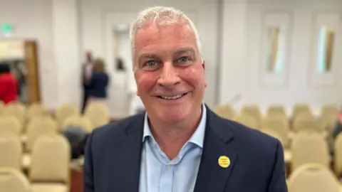 Deputy Andrew Niles - A man with short grey hair and blue eyes smiling at the camera. He is wearing a light blue shirt and a navy blue blazer.