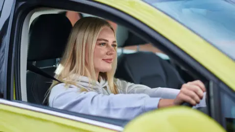 A young blonde woman is driving a yellow car. She wears a grey hoody and has a slight smile on her face