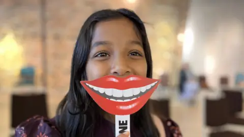 A girl holding up a mask of a huge red-lipped mouth and bright, white teeth in front of her face