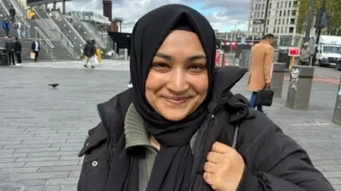 A woman in a hijab and black coat stands outside.