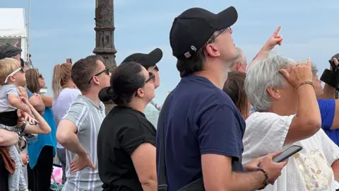 A crowd of people all look up into the sky with some pointing or covering their eyes from the sun as they watch planes fly over. The sky behind them is bright blue.