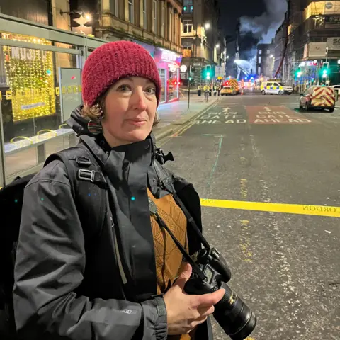 A woman in a red wollen hat and a black waterproof jacket. She has a camera hanging around her neck. She is standing in a city road and there is an emergency response to a fire in a building in the background. 