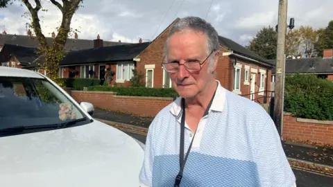 David Kelsall outside his own, standing in front of his white electric car, wearing glasses and a blue polo t-shirt 