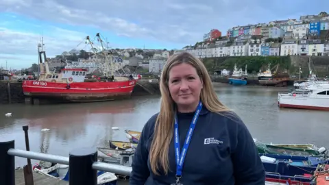 A woman standing in a harbour. The water is behind her. It is low tide. Boats are stationery on the water. She is smiling at the camera. 