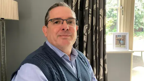 Councillor Craig Warhurst is sat down on a living room, wearing a blue shirt and sleeveless blue jumper.