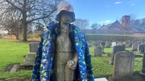 The stone memorial statue of a boy holding a ball. It has been dressed in a blue coat and a beige hat with a poppy on it and beads around the neck. Other gravestones are in the background and a church is set behind a green bush and a large tree.