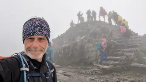 Andrew Lamb Andrew Lamb in selfie taken at top of mountain in cloudy conditions with people behind walking up to the very top on steps in mountain gear