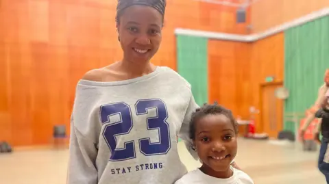 Simon Thake A woman with a grey, baggy sweatshirt smiles, with her arm around a young girl in a white t-shirt