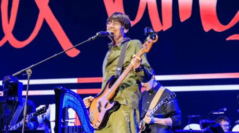 PA Media Alex James during the Alex James Britpop Classical set at Feastival 2025. He is seen on stage with a guitar and an orchestra behind him