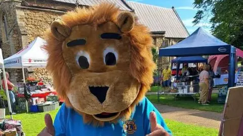 WISBECH LIONS A person wears a big lion's head and is giving a thumbs up to the camera. The lion has a blue T-shirt on and behind them are various stalls selling goods surrounding a church. 