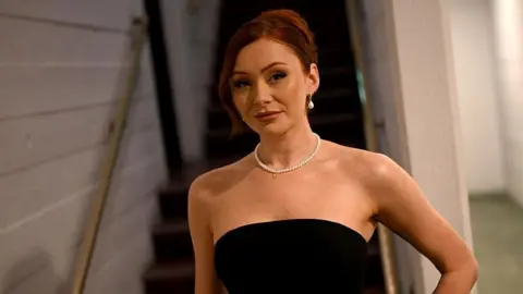 Getty Images Elz is looking straight ahead at the camera standing at the bottom of some stairs. She has her red hair tied up and is wearing a black strapless dress. She has a pearl necklace on and pearl earrings. 
