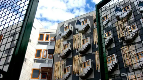 PA Media Modern building with geometric design, featuring protruding box-like windows framed in wood and decorative vertical wooden rods on a gray façade, viewed through two metal fences under a partly cloudy sky.