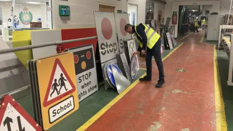 Jo Burn/BBC The Royal British Legion Industries building where veterans currently work. An employee can be seen touching a road sign, behind which is a number of other signs - including a stop sign for a school, and a stop sign when lights show.