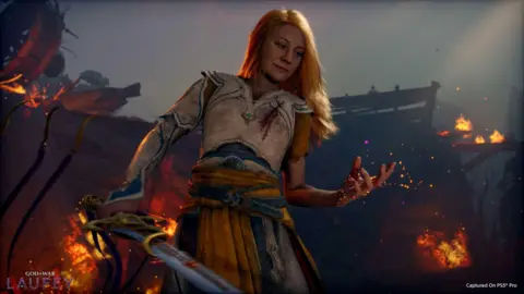 Sony Laufey is holding out her hand and looking at it, behind her things are on fire. She has long blond hair and silver armour on, she is holding a sword. 