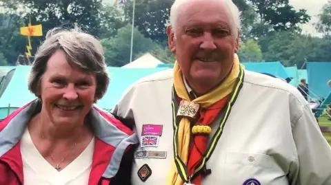 Geoff Pinney A head and shoulder shot of an older man standing with his arm around an older woman and smiling at the camera. The man is wearing a scout uniform including a neck scarf and a khaki shirt with badges. The woman is wearing a red waterproof jacket and a cream jumper. In the background can be seen a campsite of green tents and flags.