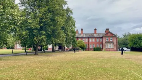 An ornate building with a large field in front of it as well as a playing field and trees. There is a row of trees to the left hand side of the image, obscuring part of the building. Two people in black clothing are standing in front of the building.
