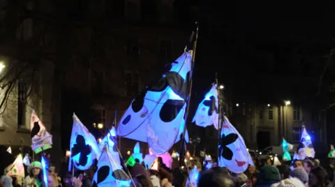 Maidstone Borough Council A dark image of Maidstone town centre with brightly lit lanterns being carried on sticks. The lanterns are white with black and red designs, they are being carried by people. 