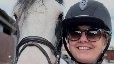 Zaphira Cormack A woman wearing a horse-riding helmet and sunglasses is smiling, posing next to a white horse.