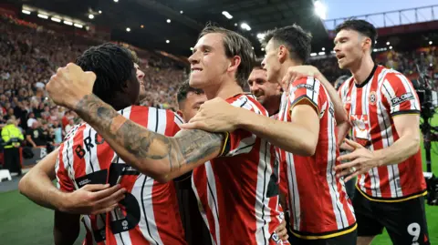 Sheffield United players celebrate a goal. Several players are embracing against the backdrop of a full floodlit stadium.