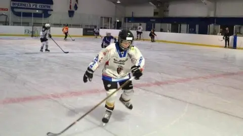 Ice hockey players skate across the ice rink inside the centre. The players are wearing helmets and padding.