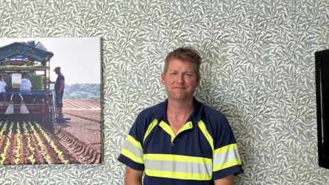 A man in a blue and yellow polo shirt stands in a room in front of wallpaper with green leaves on it. A photo of a farmer in a crop field is next to him.
