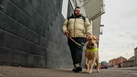 Jon Attenborough, a 36-year-old man with dark hair and a beard wearing a puffer jacket, walks his guide dog Sam outside Dundee United's Tannadice