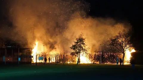 Dan Jessup Large flames coming from hedges and trees surrounding football pitches. The sky is dark and firefighters and members of the public are standing nearby.