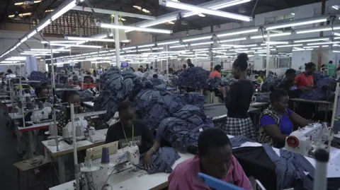 A wide shot of a busy factory floor of the garment factory. Workers at sewing machines and piles of finished items can be seen.