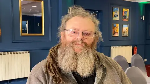 Lance Vaudin He has long grey hair and a grey beard. He is wearing glasses and a grey hooded jumper. 