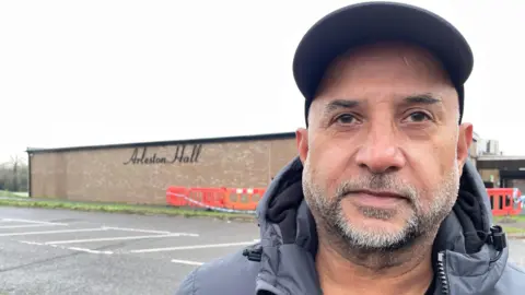 Sohan Perdesi is pictured wearing a cap. He is stood in front of a building with the words Arleston Hall visible.