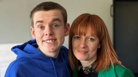 Caleb standing next to his mother, Alma White, as they pose for a photo.   Caleb is a teenager with short, fair hair and blue eyes.  He is wearing a blue hooded top.  Alma has shoulder-length ginger hair with a fringe.  She is wearing a bright green coat over a black top with a patterned collar. 