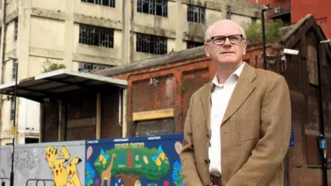 Ipswich Borough Council Neil McDonald wearing a brown jacket and glasses. He is standing in front of some graffiti on a derelict building and looking off in the distance. 