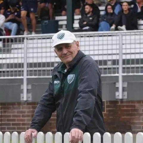 Randwick Rugby Club Peter Meagher leans on a low white picket fence. He wears a grey jacket with a green rugby logo, and a white sun hat with the same logo. Behind him, spectators are sat in a set of white stands.