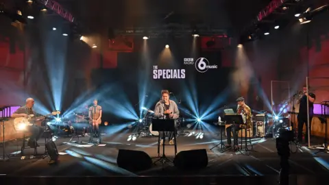 Terry Hall and The Specials are seen in a BBC studio with BBC Radio 6 Music and The Specials branding in the background. The studio is lit with blue uplights.