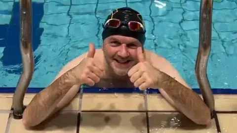 Michael Coombes, a man wearing a swimming cap and leaning on the edge of an indoor pool, gives a thumbs up.