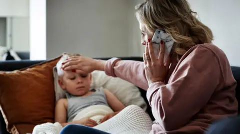 Getty Images Woman and child on a sofa. The woman is on the phone while touching the forehead of the child, who looks ill