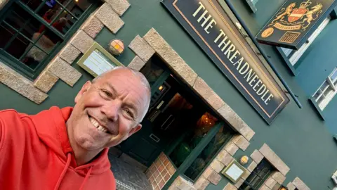 David Bennett David Bennett wearing a bright orange hoodie while taking a selfie smiling outside a pub
