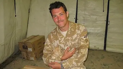Handout Andrew Borthwick wearing a khaki camouflage military uniform, sitting inside a canvas tent while on deployment in Afghanistan. He has dark hair, brown eyes and a thin moustache and is smiling at the camera with his arms folded in front of him.