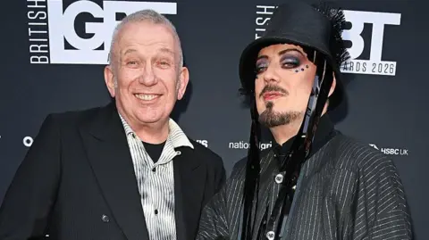 Getty Images Jean Paul Gaultier and Boy George stand in front of a black wall with the logo 'British LGBT Awards' on it. Jean is wearing a striped white and grey shirt with a black blazer and is smiling. Boy George is wearing a pinstripe jacket and a black bucket-style hat with tassles. He has facial hair and gems around his eyes. 