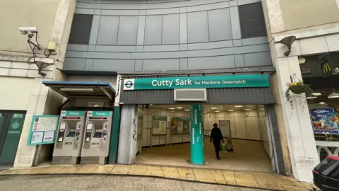 Front of Cutty Sark station in Greenwich. The picture shows two ticket mchines and the entrance hall during daylight, with one person walking in